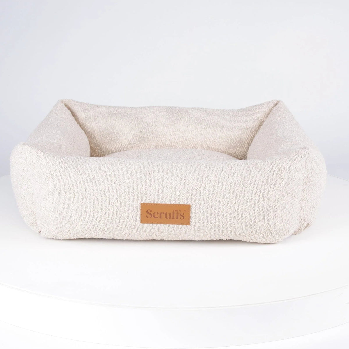 A cream-colored sheepskin pet bed with a small brown leather 'Scruffs' brand patch, displayed on a white circular platform against a white background.