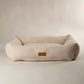 A plush, low-profile pet bed in a textured cream bouclé fabric, featuring a small brown leather brand patch on the front side. The bed is photographed on a seamless, light beige studio background.