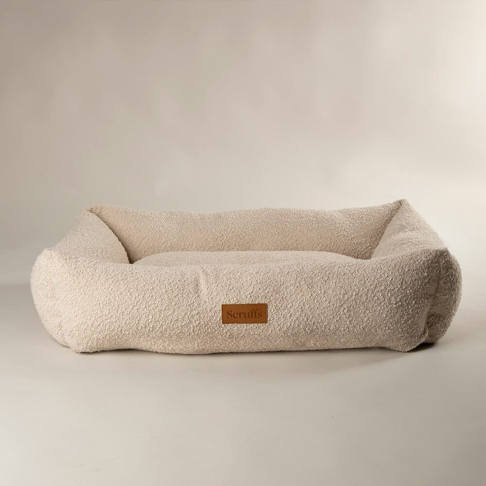 A plush, low-profile pet bed in a textured cream bouclé fabric, featuring a small brown leather brand patch on the front side. The bed is photographed on a seamless, light beige studio background.