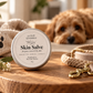 A round tin of poochiella KINDRED Skin Salve sits on a wooden surface in a warm, cozy setting with a fluffy dog in the background, alongside a knit blanket with a paw print, flowers, and a rope leash.