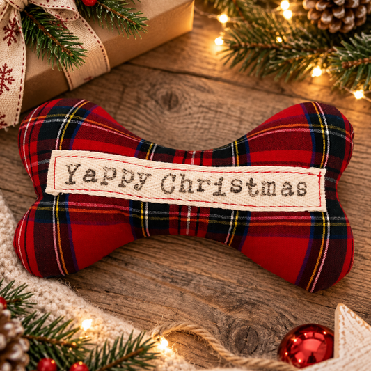 A bone-shaped Christmas dog toy made of red tartan plaid fabric with a cream embroidered label reading 'Happy Christmas,' styled on a rustic wooden surface with holiday greenery, lights, pinecones, and gift decor.