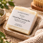 A bar of poochella KINDRED Natural Dog Cleansing Bar sits in a speckled stone dish on a textured beige surface, surrounded by soft home props including a knit blanket, green leaves, and a wooden brush.