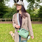 Woman holding a green crossbody bag in a park with trees and grass in the background