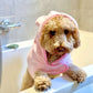 Dog wrapped in a pink towel sitting on a bathtub rim.