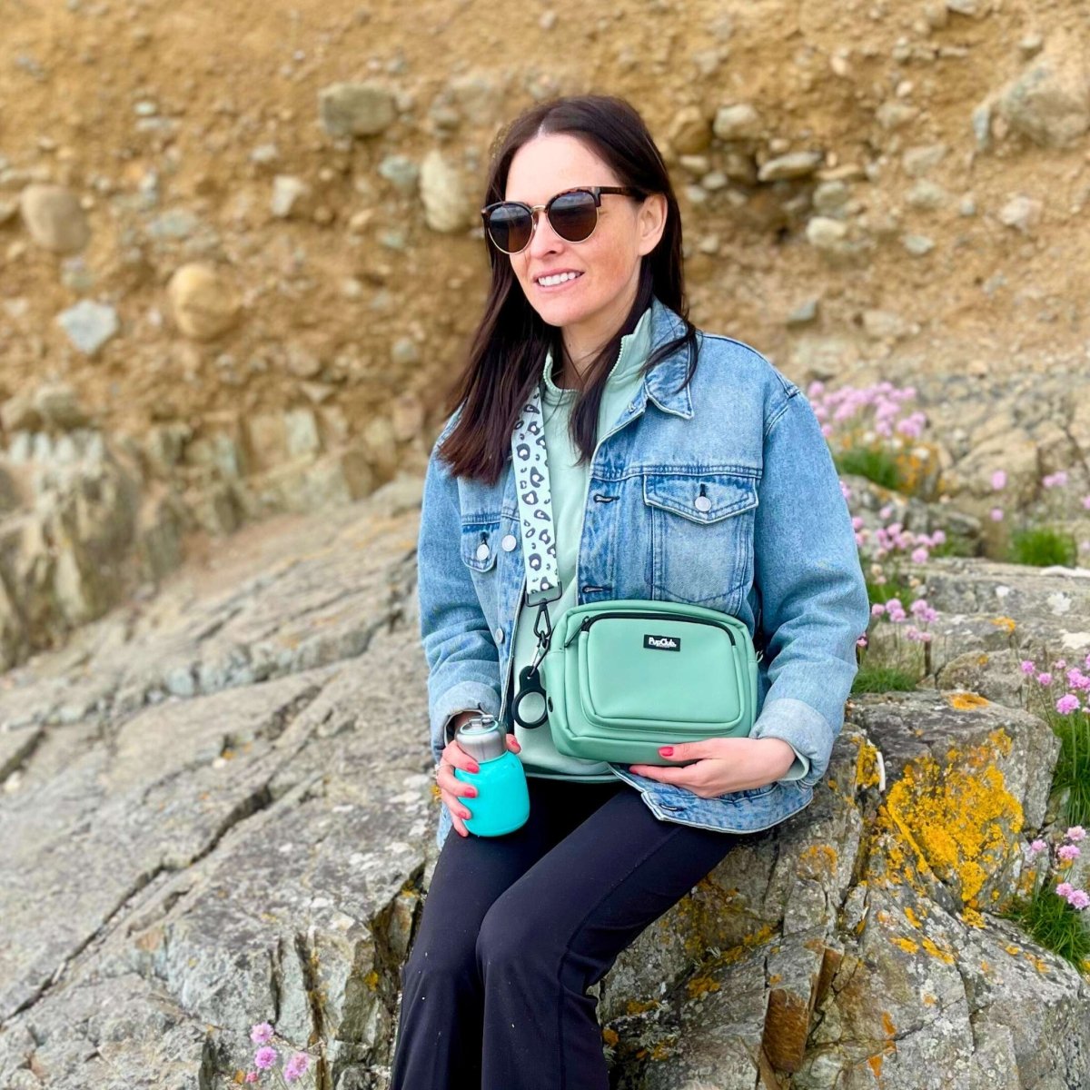 Woman holding a green crossbody bag on rocky terrain with flowers.