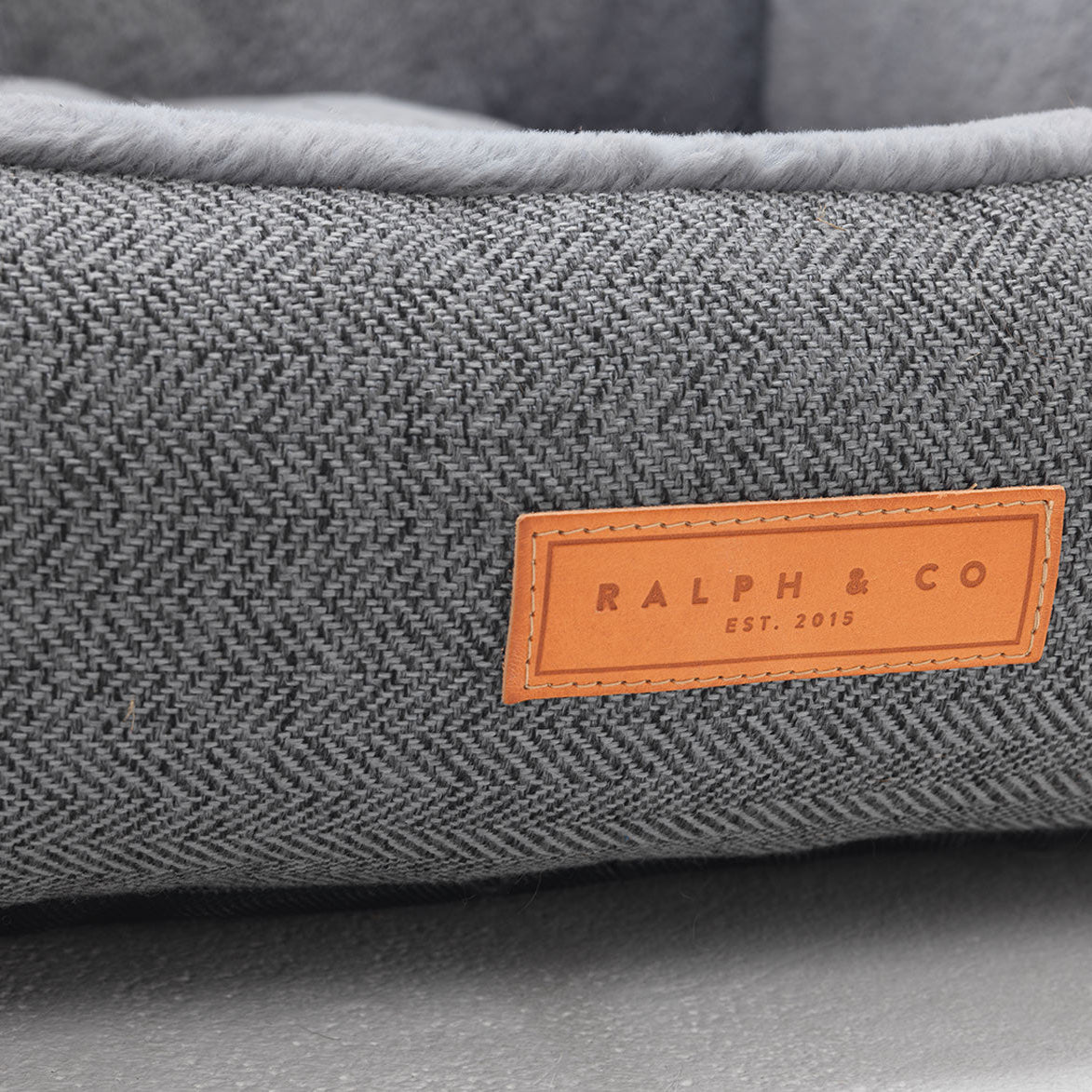 Gray textured pet bed with a brown leather label reading 'Ralph & Co' on a gray background.