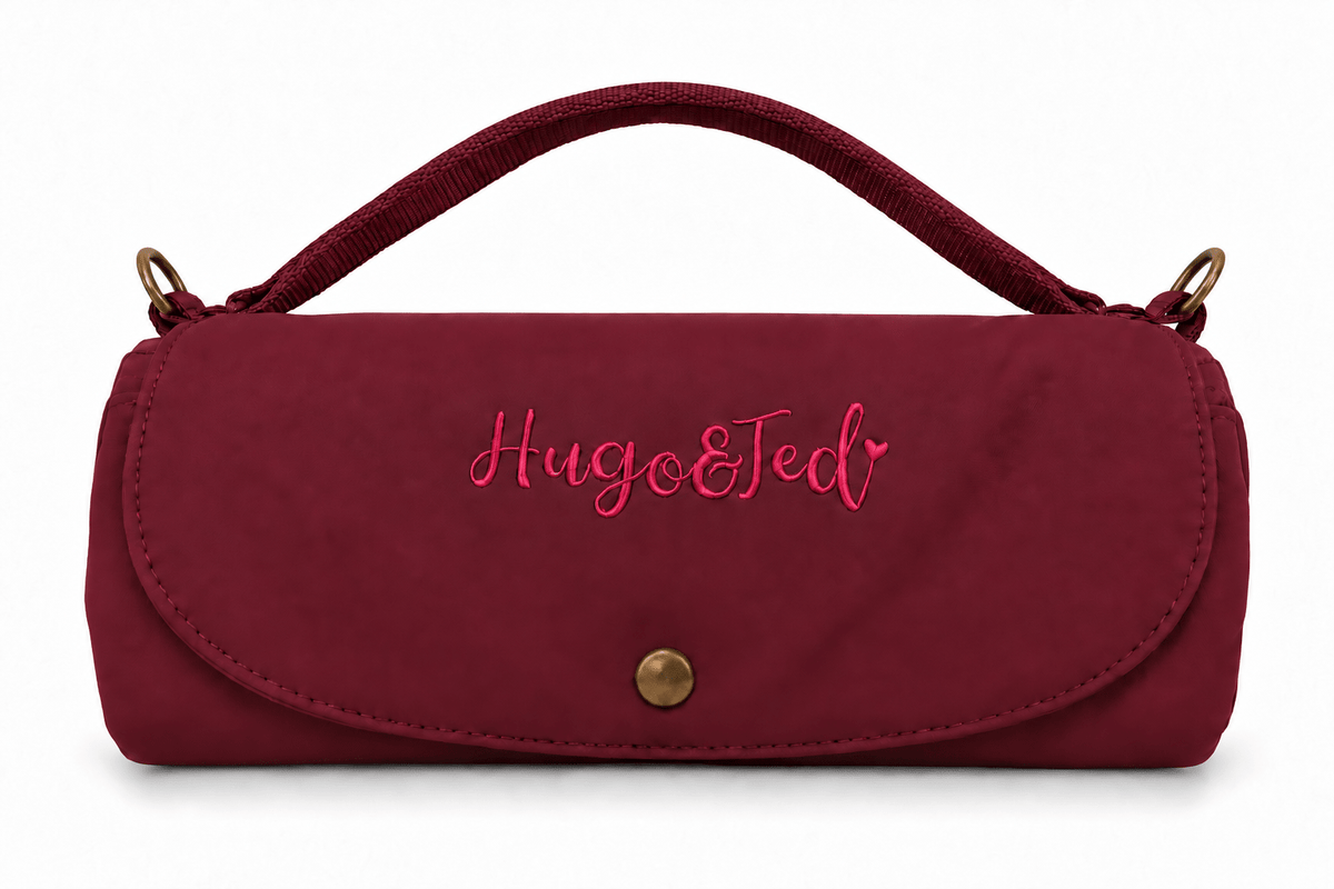 A burgundy nylon clutch bag with a top handle, featuring the embroidered pink text 'Hugo&Ted' on the front flap and a gold snap button closure.