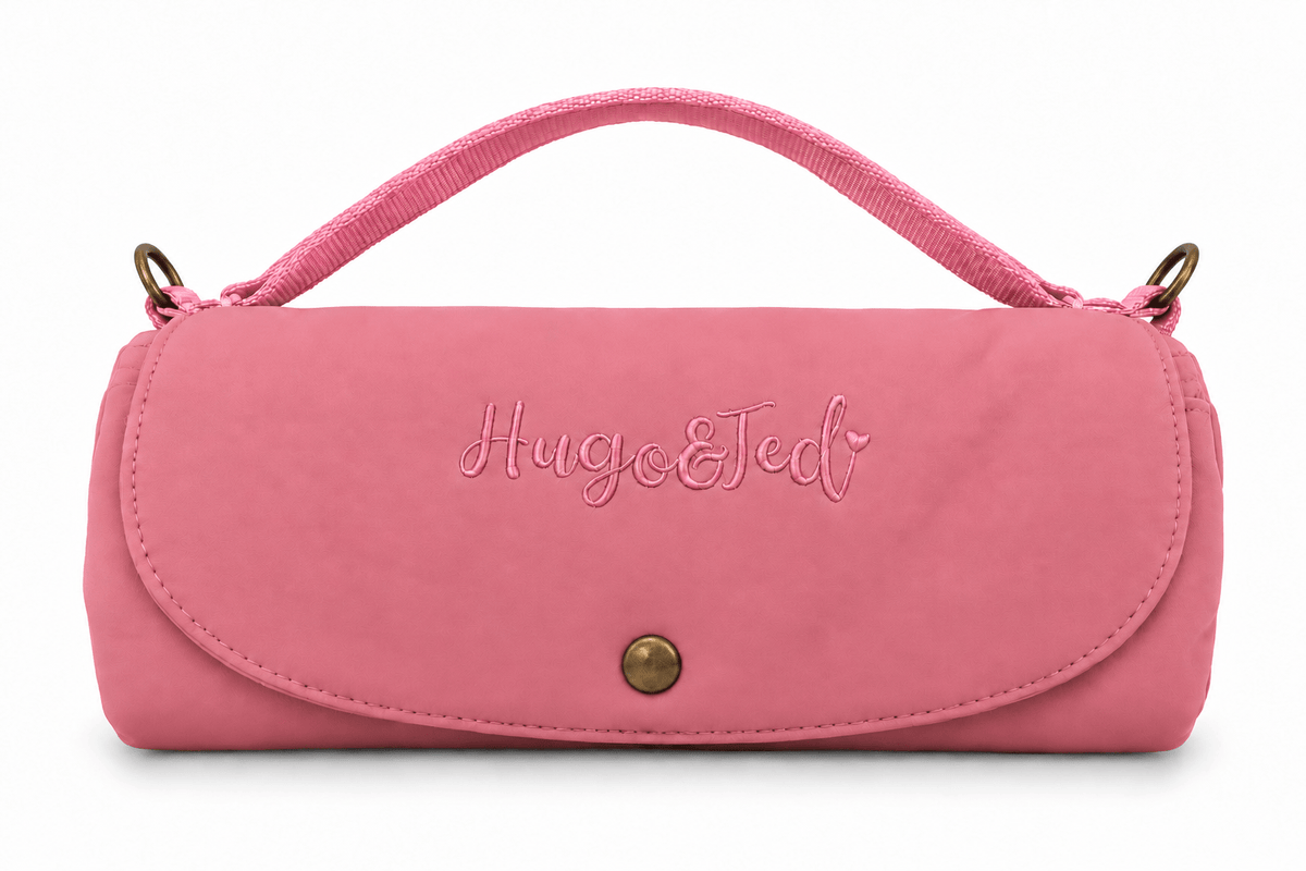 A pink  handbag with a top handle, featuring the brand name 'Hugo & Ted' embroidered on the front flap and a gold snap button closure, set against a plain white background.