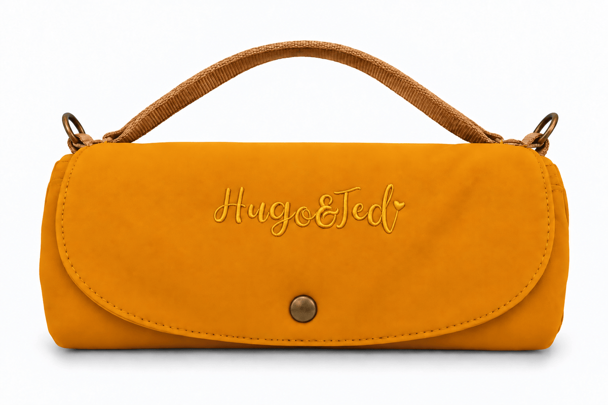 A mustard yellow faux suede clutch bag with a brown woven top handle, featuring the embroidered brand name 'Hugo & Ted' on the front flap and a gold snap button closure.