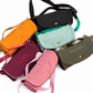 Six rolled fabric shoulder bags in different colors (orange, black, mint green, burgundy, pink, and olive green) arranged in a fan shape on a white background. Each bag features a flap closure with a snap button, a small embroidered brand name, and a matching fabric strap.