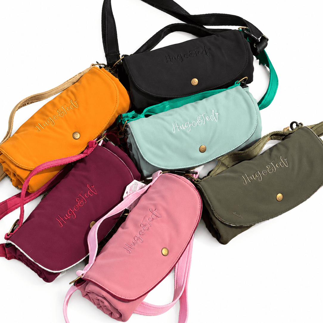 Six rolled fabric shoulder bags in different colors (orange, black, mint green, burgundy, pink, and olive green) arranged in a fan shape on a white background. Each bag features a flap closure with a snap button, a small embroidered brand name, and a matching fabric strap.