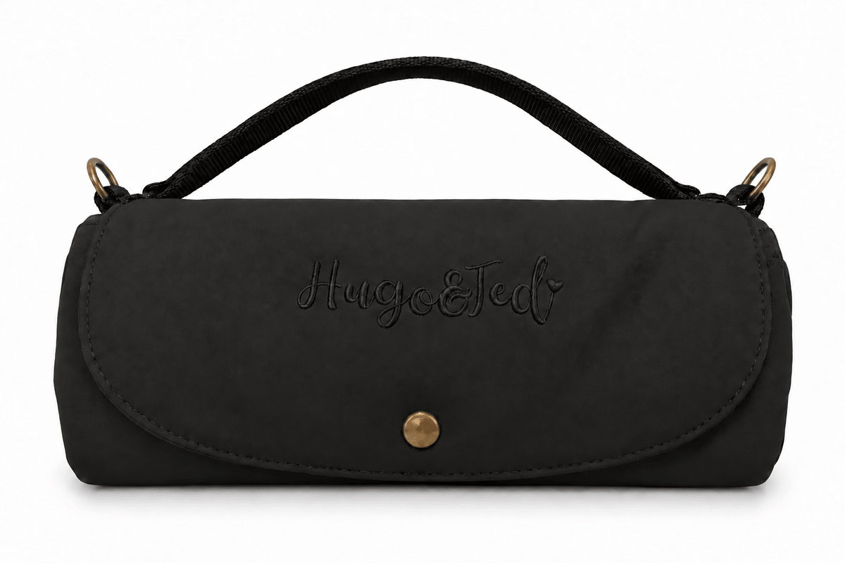 A black fabric handbag with a top handle, featuring the brand name 'Hugo & Ted' embroidered on the front flap and a gold snap button closure, set against a plain white background.