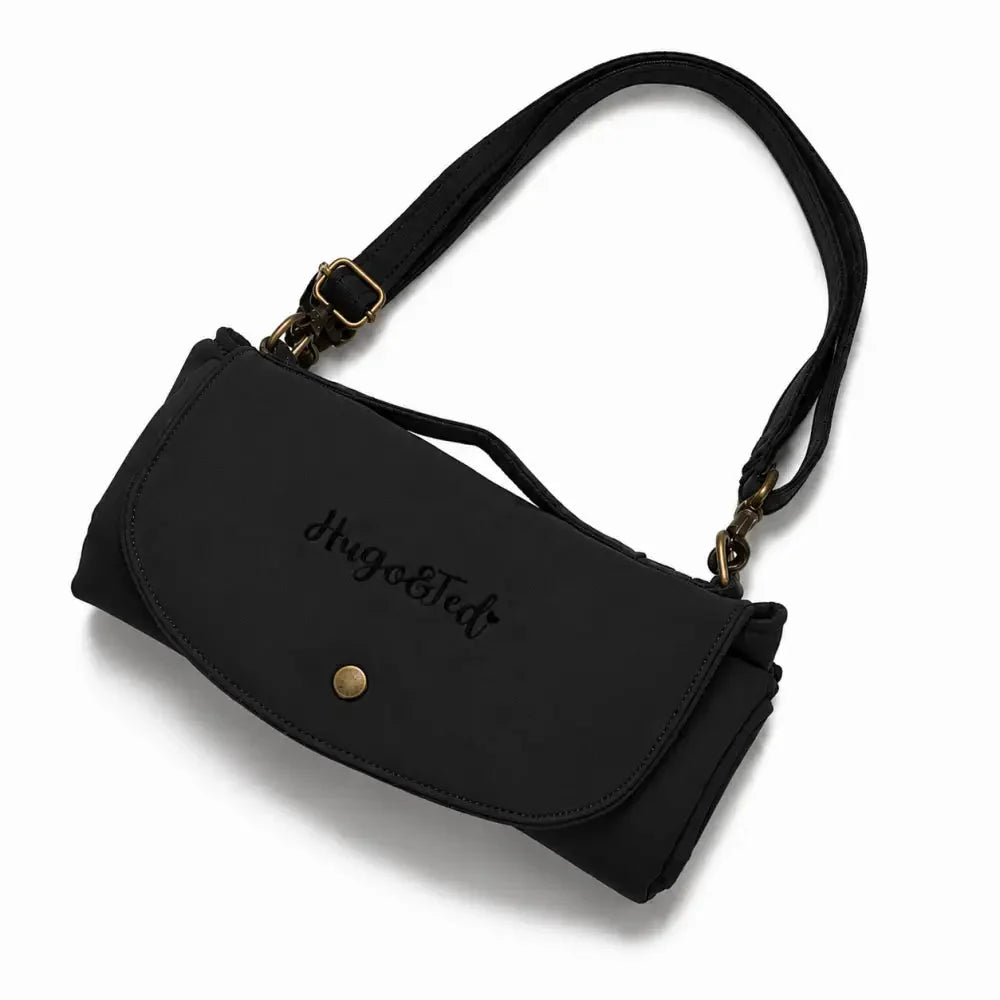 A black Hugo & Ted crossbody  folding dog travel mat bag with a curved flap, embossed logo, and gold-tone hardware, shown on a white background.