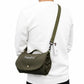 An olive green crossbody bag with a top handle and adjustable shoulder strap, worn by a person in a white sweatshirt. The bag features white embroidered text reading 'Hugo & ted' on the front flap and a small gold snap button.