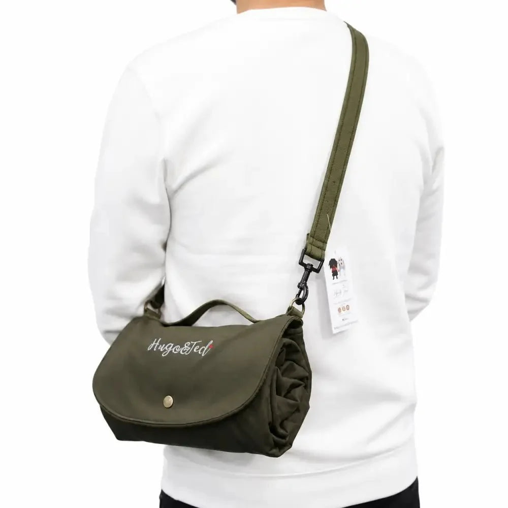 An olive green crossbody bag with a top handle and adjustable shoulder strap, worn by a person in a white sweatshirt. The bag features white embroidered text reading 'Hugo & ted' on the front flap and a small gold snap button.