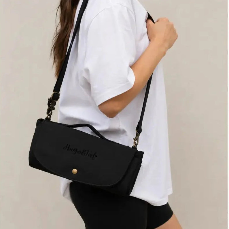 A woman wearing a white t-shirt and black bike shorts carries a black crossbody bag with a top handle and a gold snap button. The bag features the script logo 'Hugo & Ted' on the front flap.